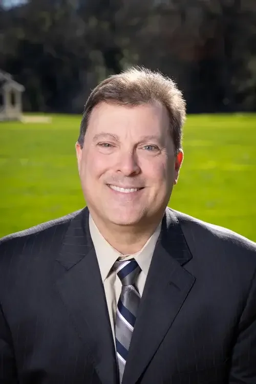 Portrait of a middle-aged man smiling outdoors with a green lawn and trees in the background, wearing a suit and tie.