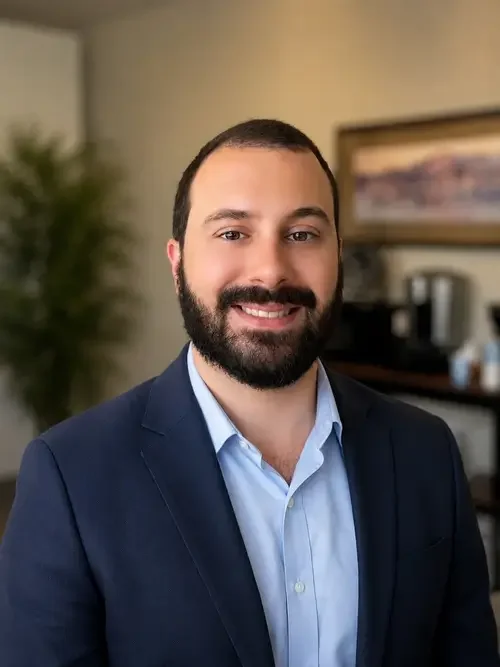 A smiling man with a beard, wearing a navy blazer and light blue dress shirt, posed indoors with a blurred background of a plant and framed picture.