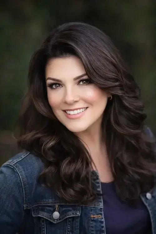 A smiling woman with long dark curly hair, wearing a denim jacket, outdoors with a blurred green background.