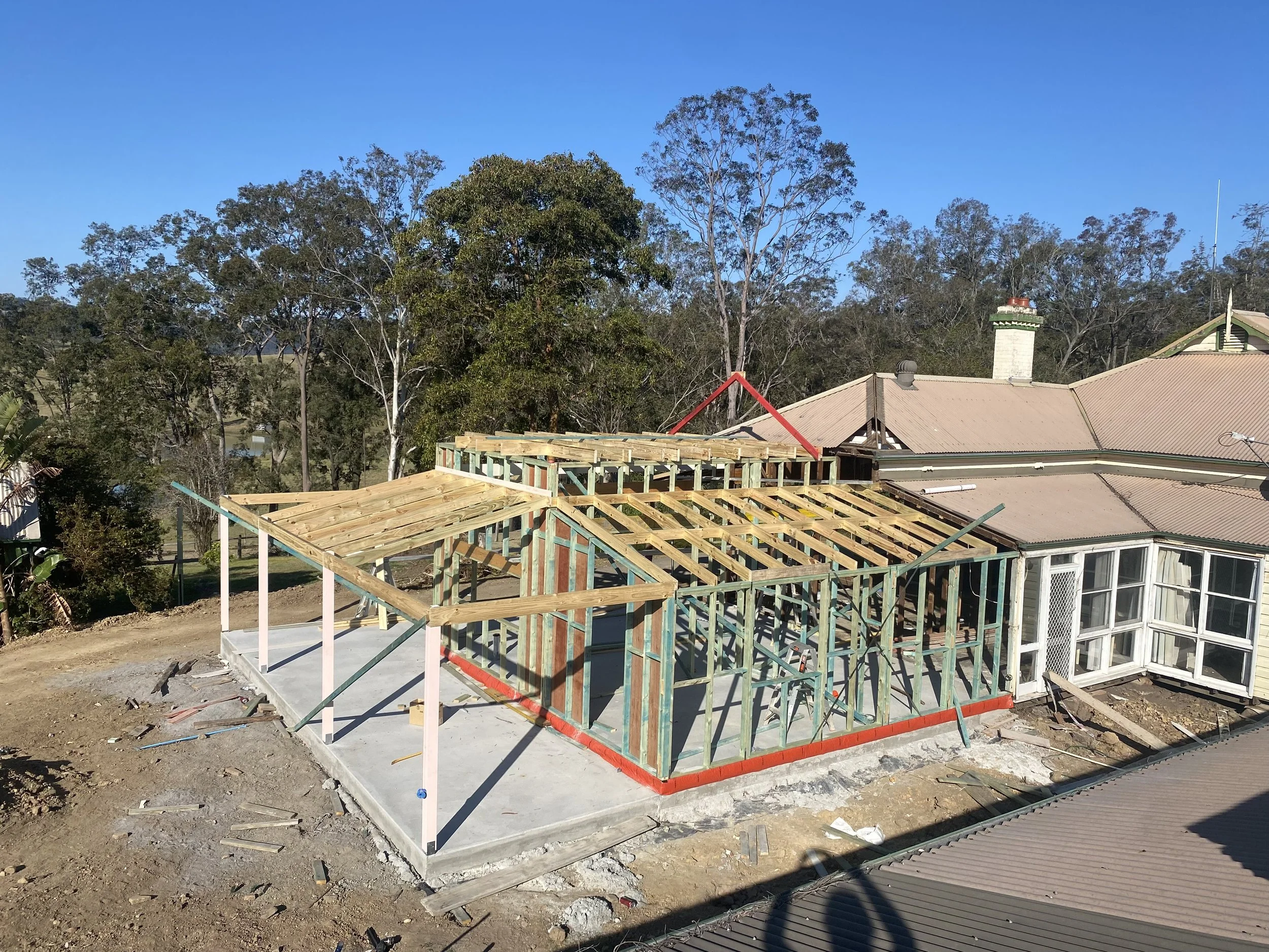 Residential house under construction with wooden framing on a concrete slab, adjacent to finished house with a tiled roof and chimney