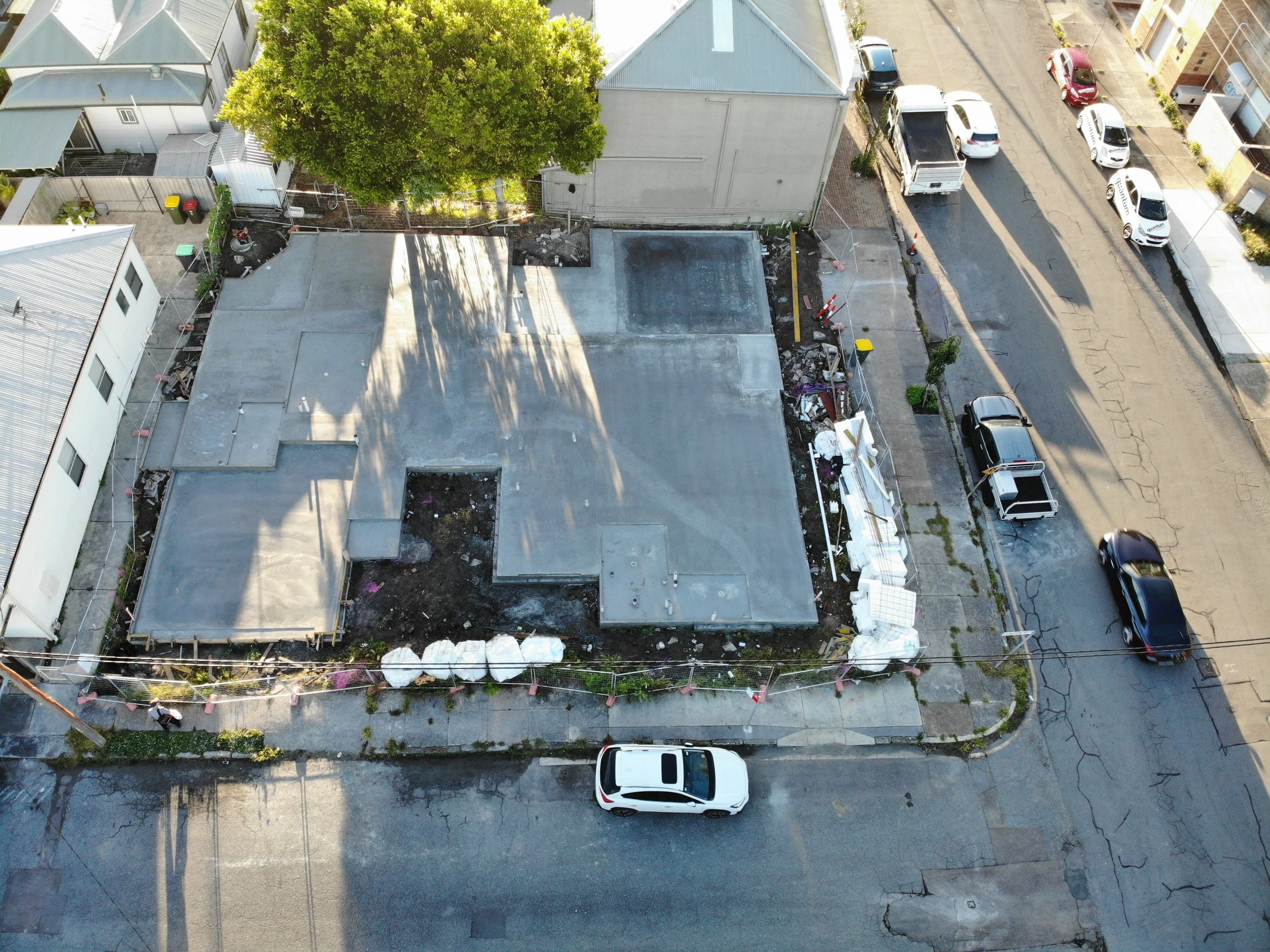 Aerial view of a construction site behind a residential area with parked cars along the street. The construction site has a new concrete foundation and surrounding fencing.