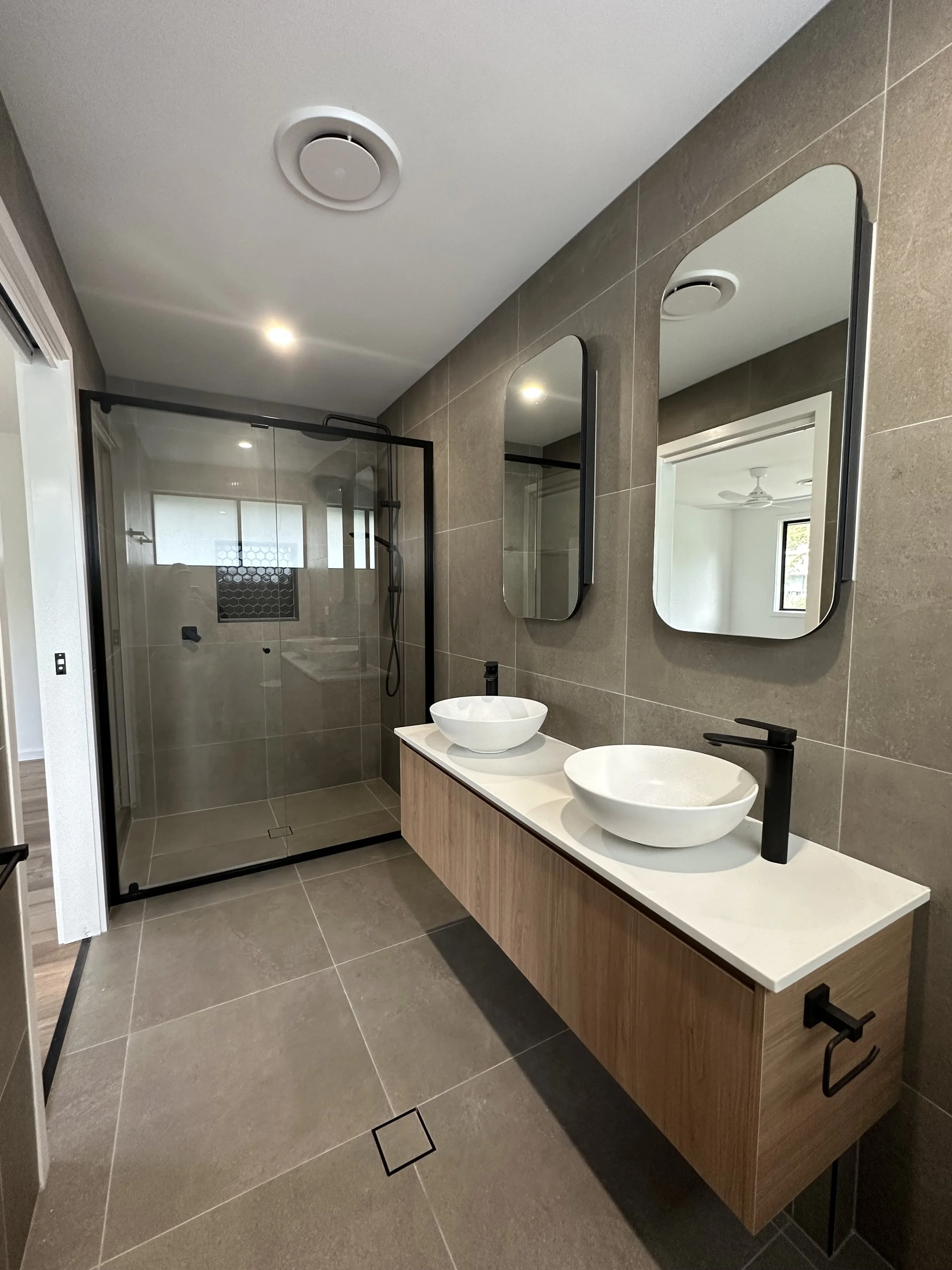 Modern bathroom with vanity featuring two vessel sinks, mirrors, and a walk-in shower with glass door and black fixtures, beige tiled walls, and gray tiled floor.