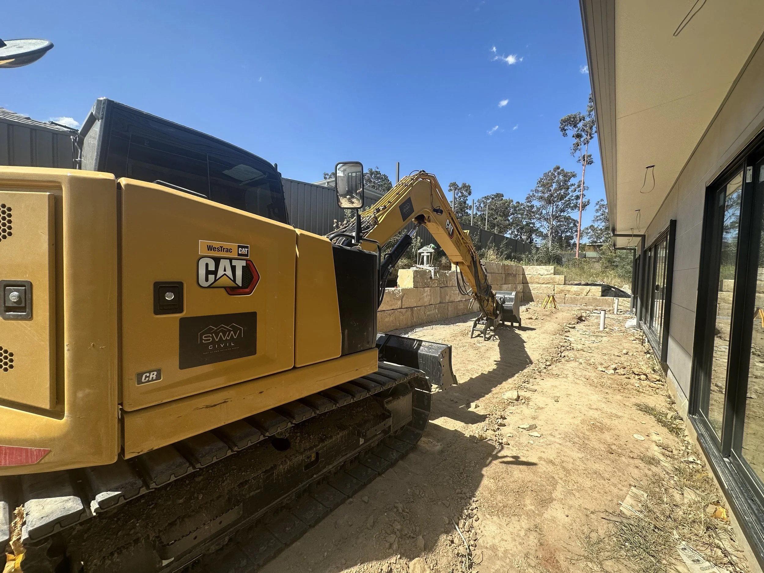 Construction site with a large yellow CAT excavator and a partially built retaining wall made of concrete blocks 