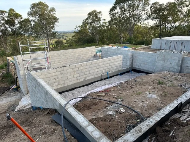 Construction site with concrete block foundation in progress, surrounded by dirt and construction tools, with trees and landscape in the background.