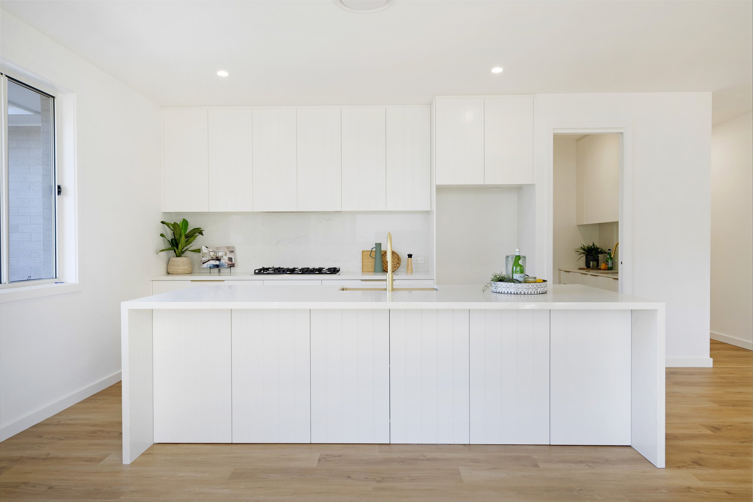 Modern kitchen with white cabinetry, a kitchen island, a gas stove, a brass faucet, and minimal decor including plants and bottles, with a window on the left and a doorway on the right.