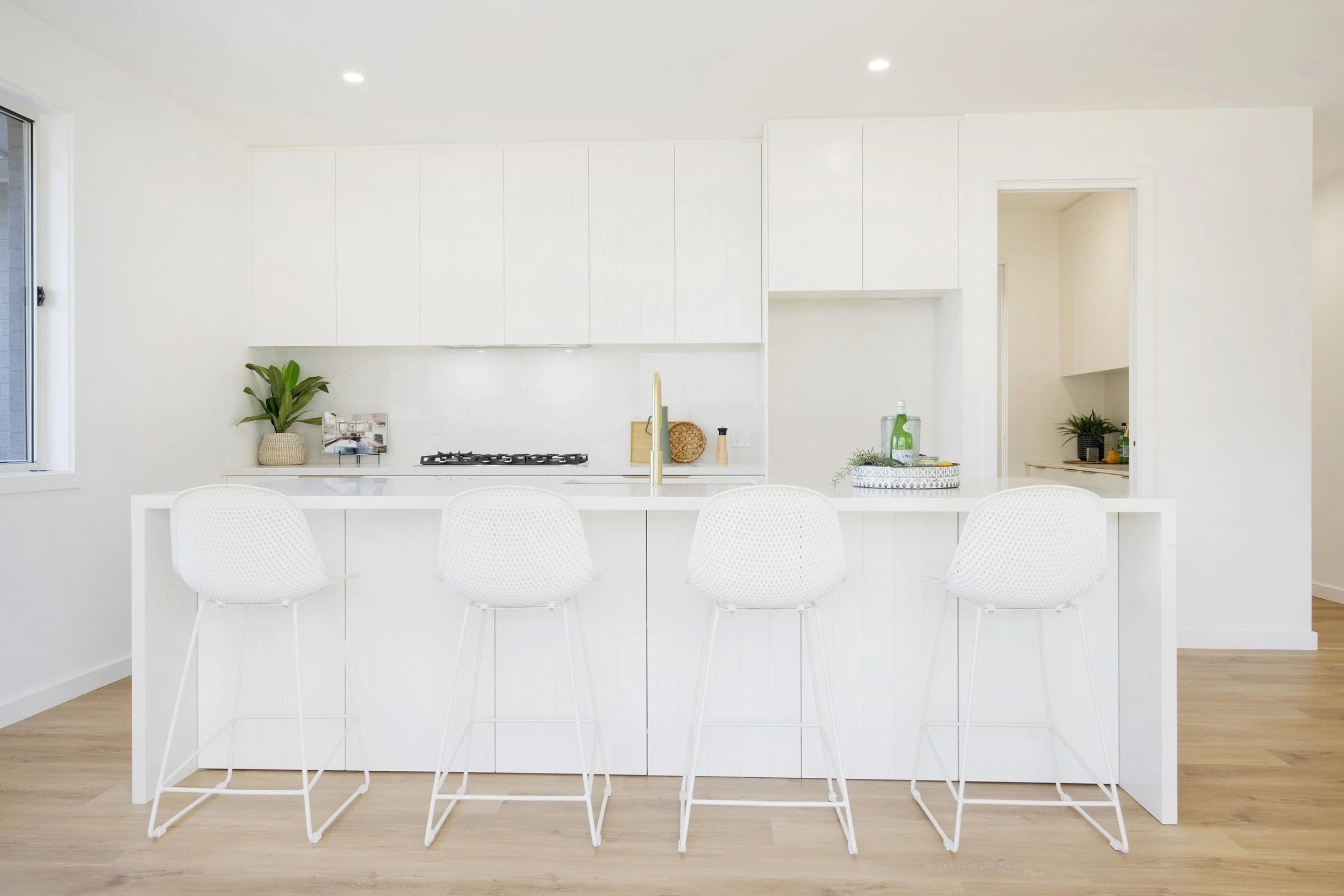 Modern white kitchen with island, bar stools, potted plant, and natural light from window, with dining area in foreground.