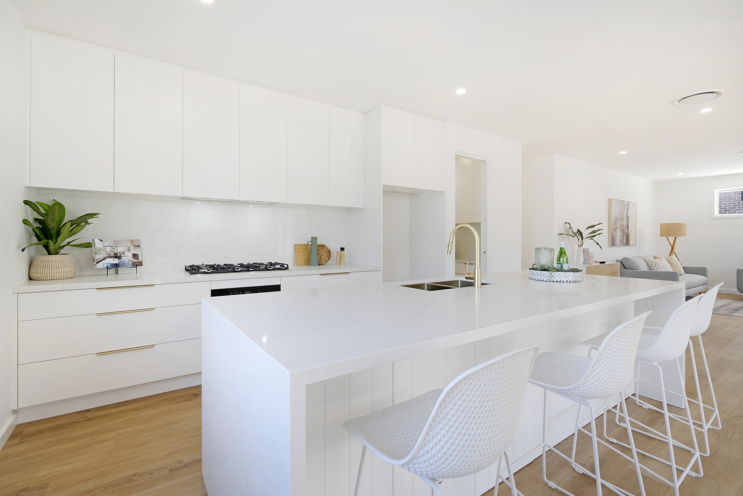 A modern, minimalist white kitchen and living room with a large island, white cabinetry, a potted plant, and white chairs.