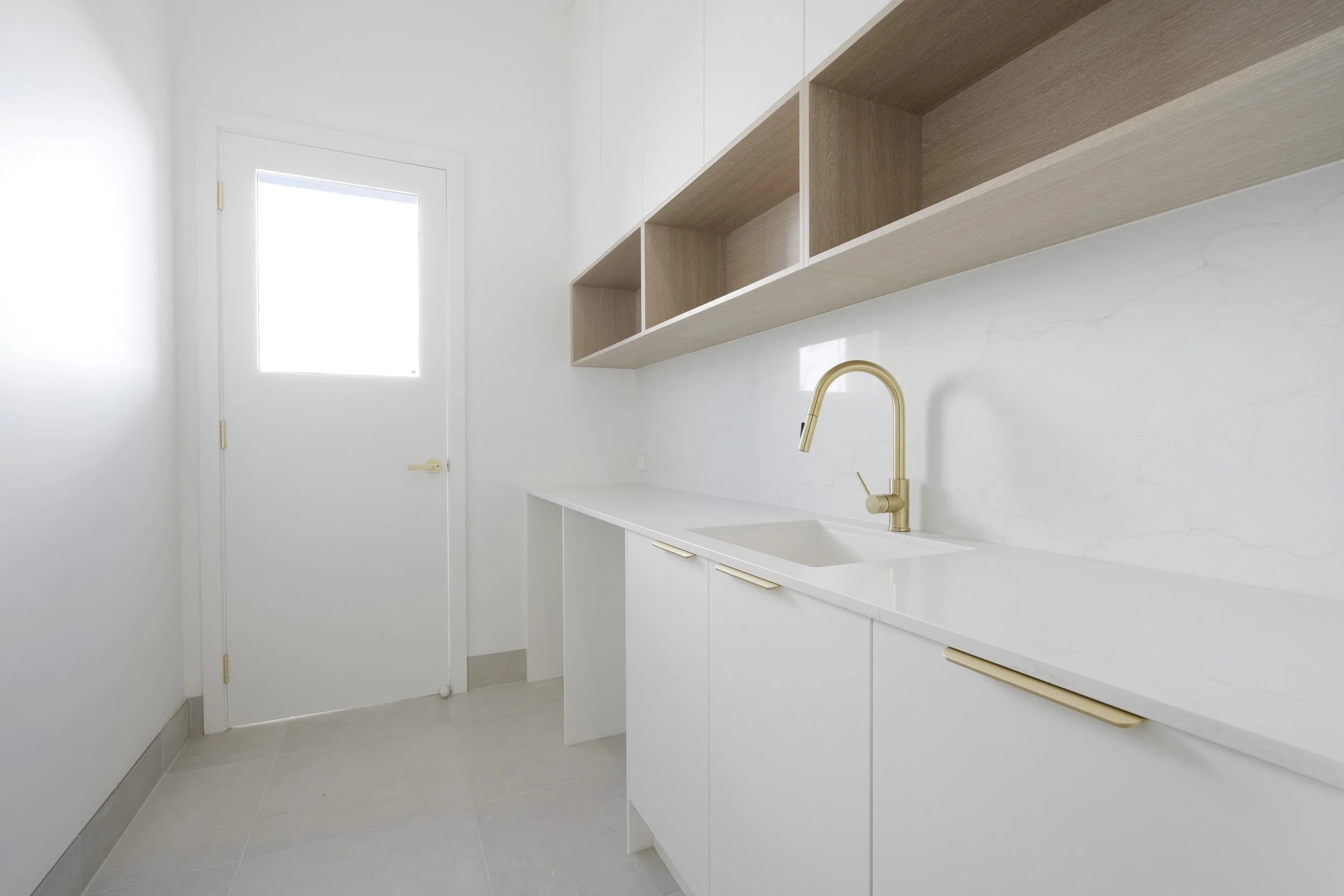 Modern minimalist kitchen with white cabinets, a gold faucet, wooden open shelves, and a window in Hunter's Valley custom built by SWW BUILDING GROUP