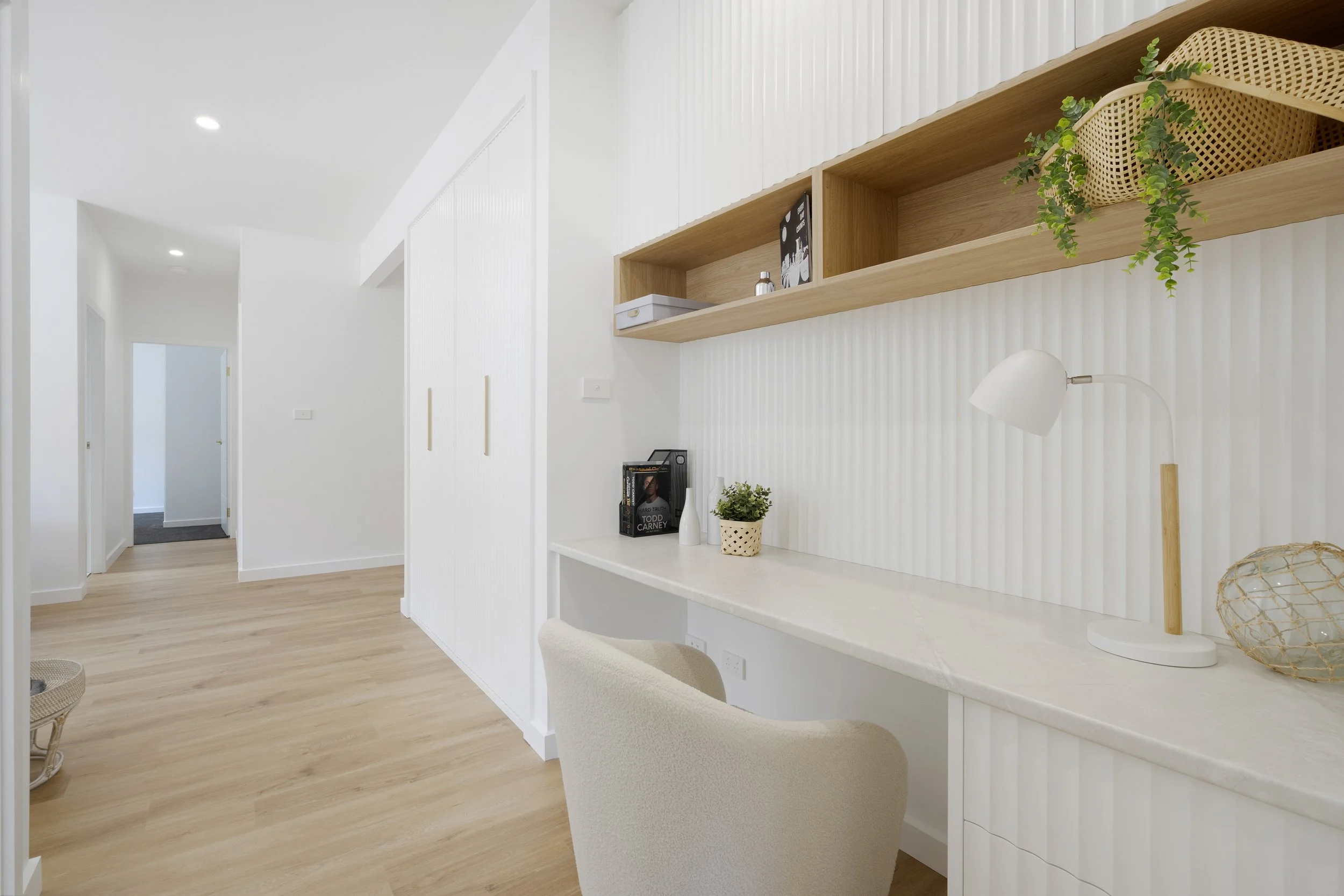 White room with wooden accents, a built-in desk with small plants and decorative items, and open wooden shelves displaying books and a plant.