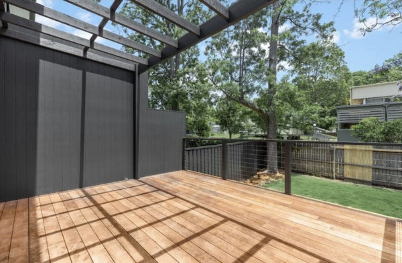 Empty outdoor deck with wooden flooring, black railing, and a partial view of trees and neighboring houses under a bright blue sky.