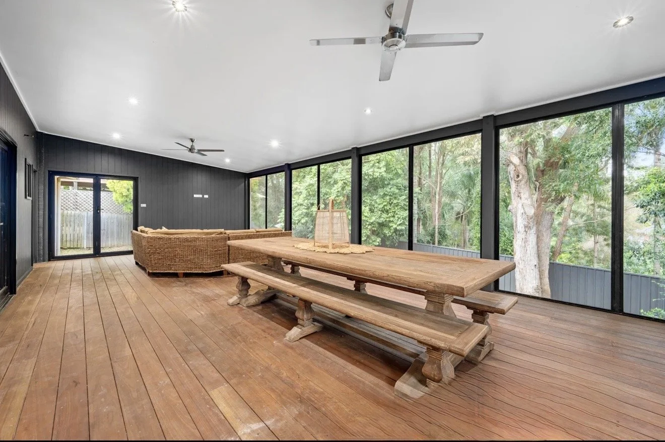 Enclosed outdoor patio with wooden floor, large wooden dining table with benches, wicker sectional sofa, ceiling fans, and large windows showing trees outside.