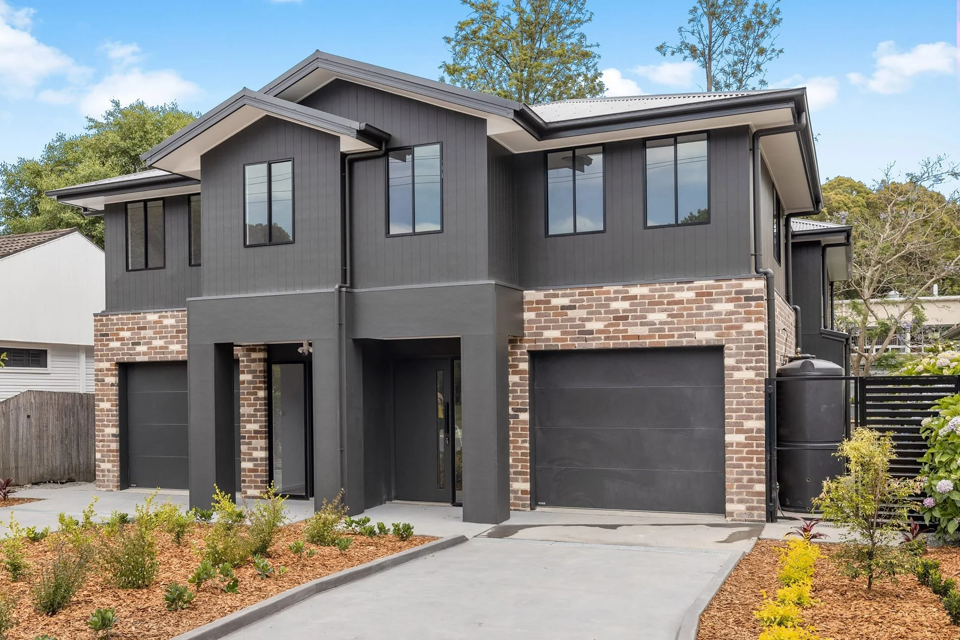 Modern two-story house with dark gray exterior, brick accents, large black-framed windows, and a black garage door. Front yard has small shrubs and a concrete driveway.