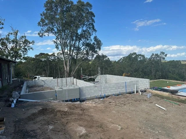 Construction site with a concrete block foundation and partial walls, surrounded by trees and blue sky.