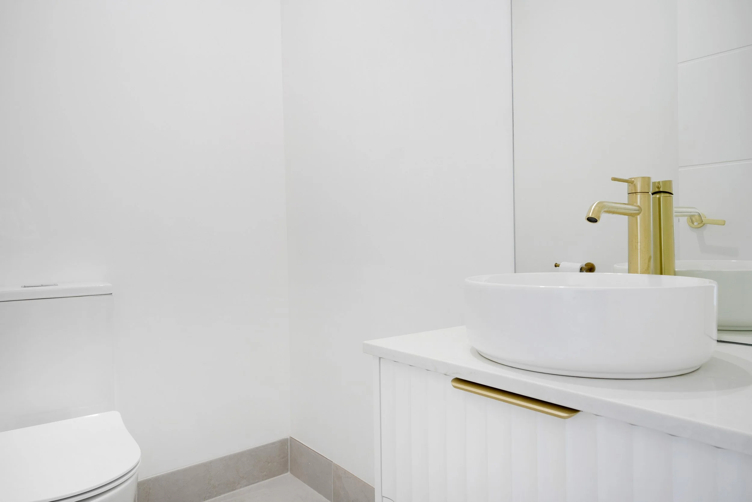 A modern bathroom with white walls, a white sink with a gold faucet, and a partial view of a toilet, with beige tiled flooring.