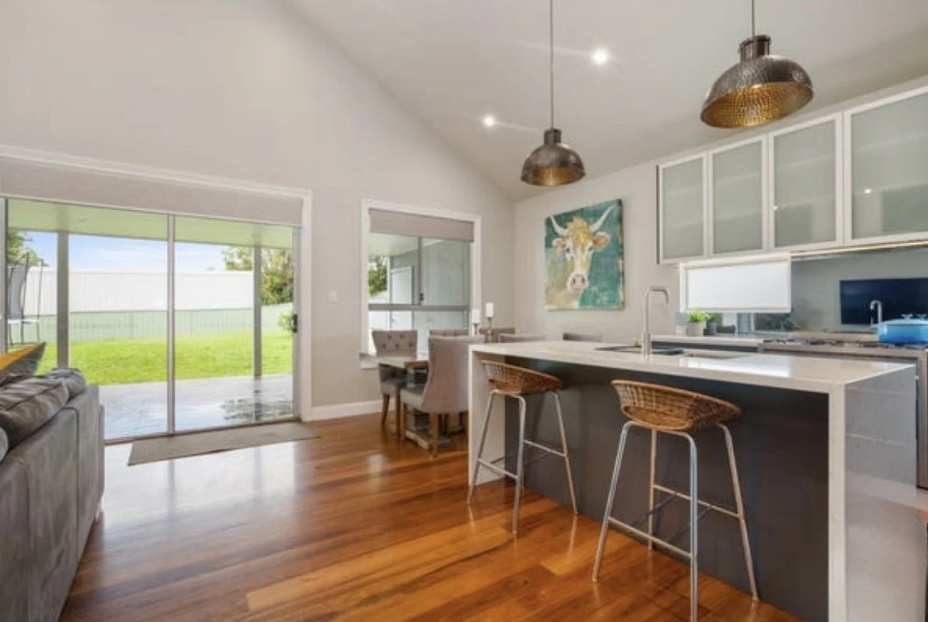 Modern kitchen with white cabinets, a large island with seating, and a painting of a cow on the wall. There are sliding glass doors leading to a backyard