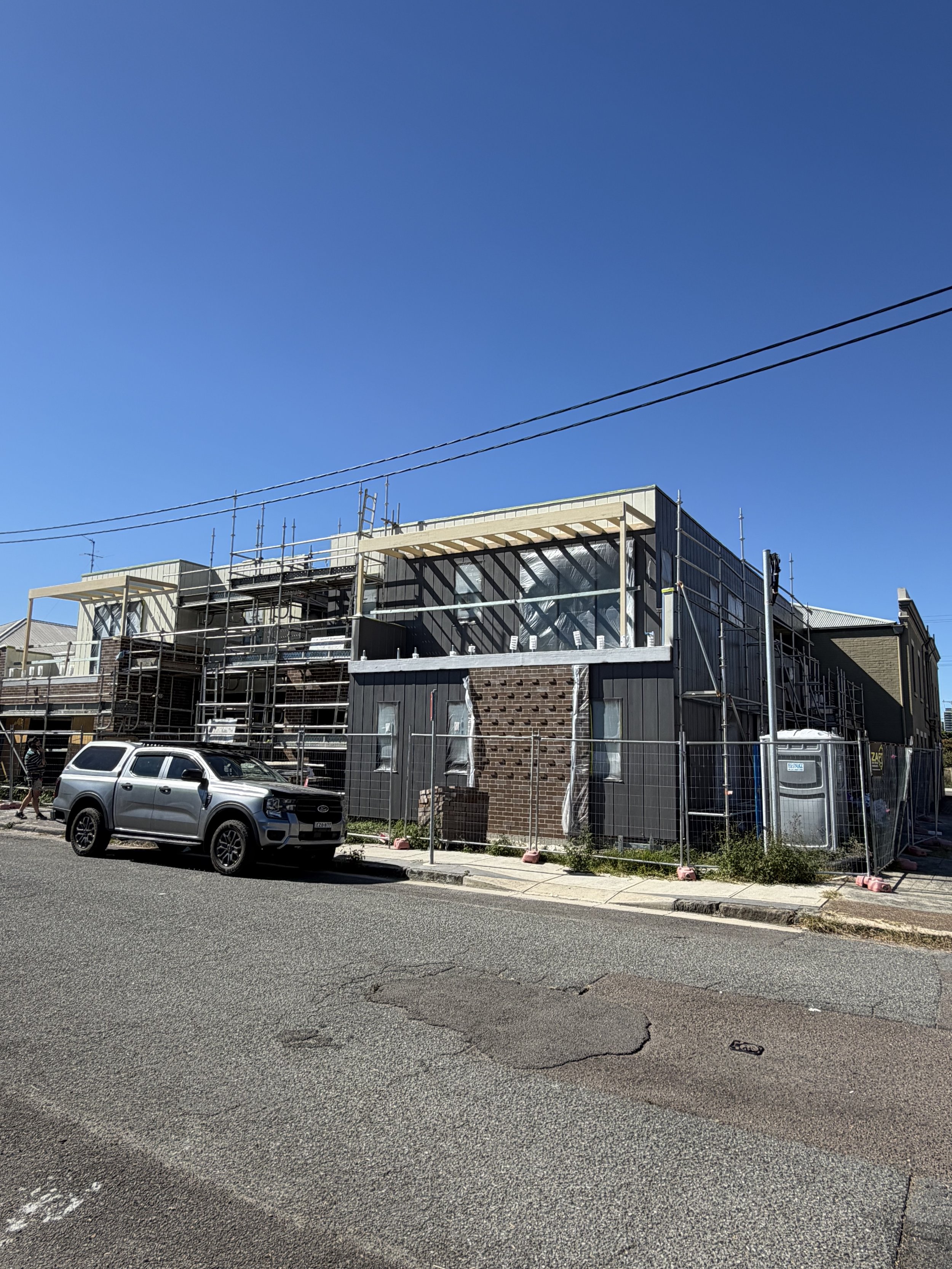 A building under construction with scaffolding, fencing, and construction materials, situated along a paved street with parked cars and a pedestrian walking beside it. The sky is clear and blue.