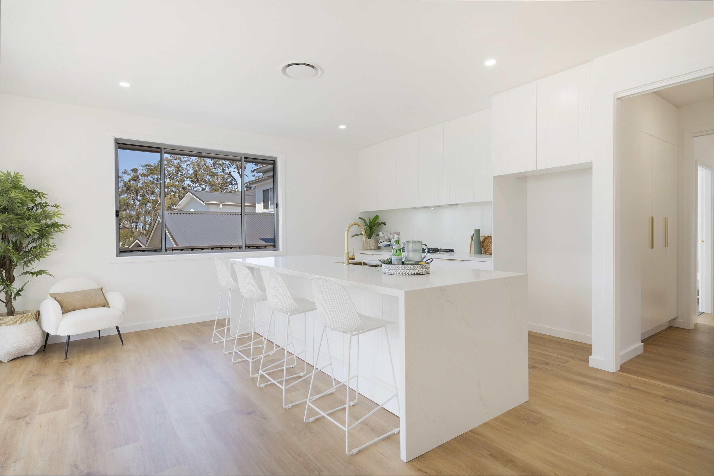 Interior view of a modern, minimalistic kitchen and living area with white walls, wooden floor, a large window showing outdoor trees, a breakfast bar in Hunter's Valley custom home built by SWW BUILDING GROUP