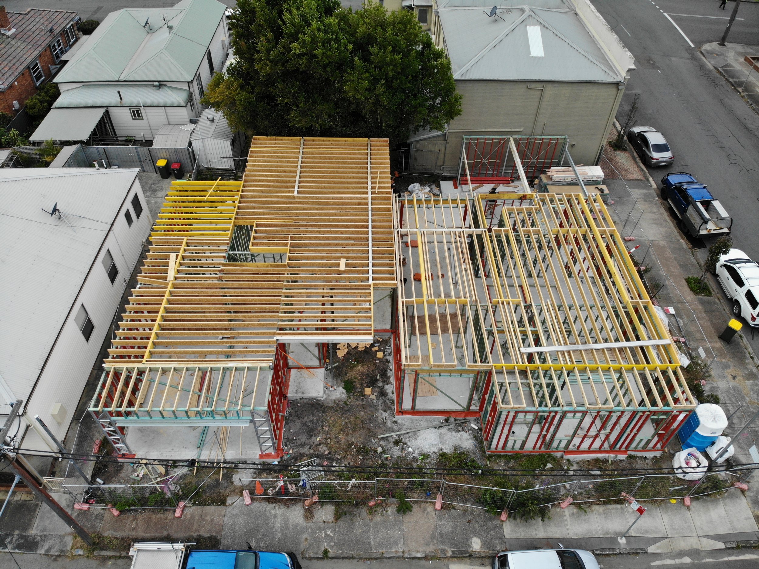 Aerial view of a construction site in an urban neighborhood with houses, parked cars, trees, and ongoing building framework with wooden and metal beams.