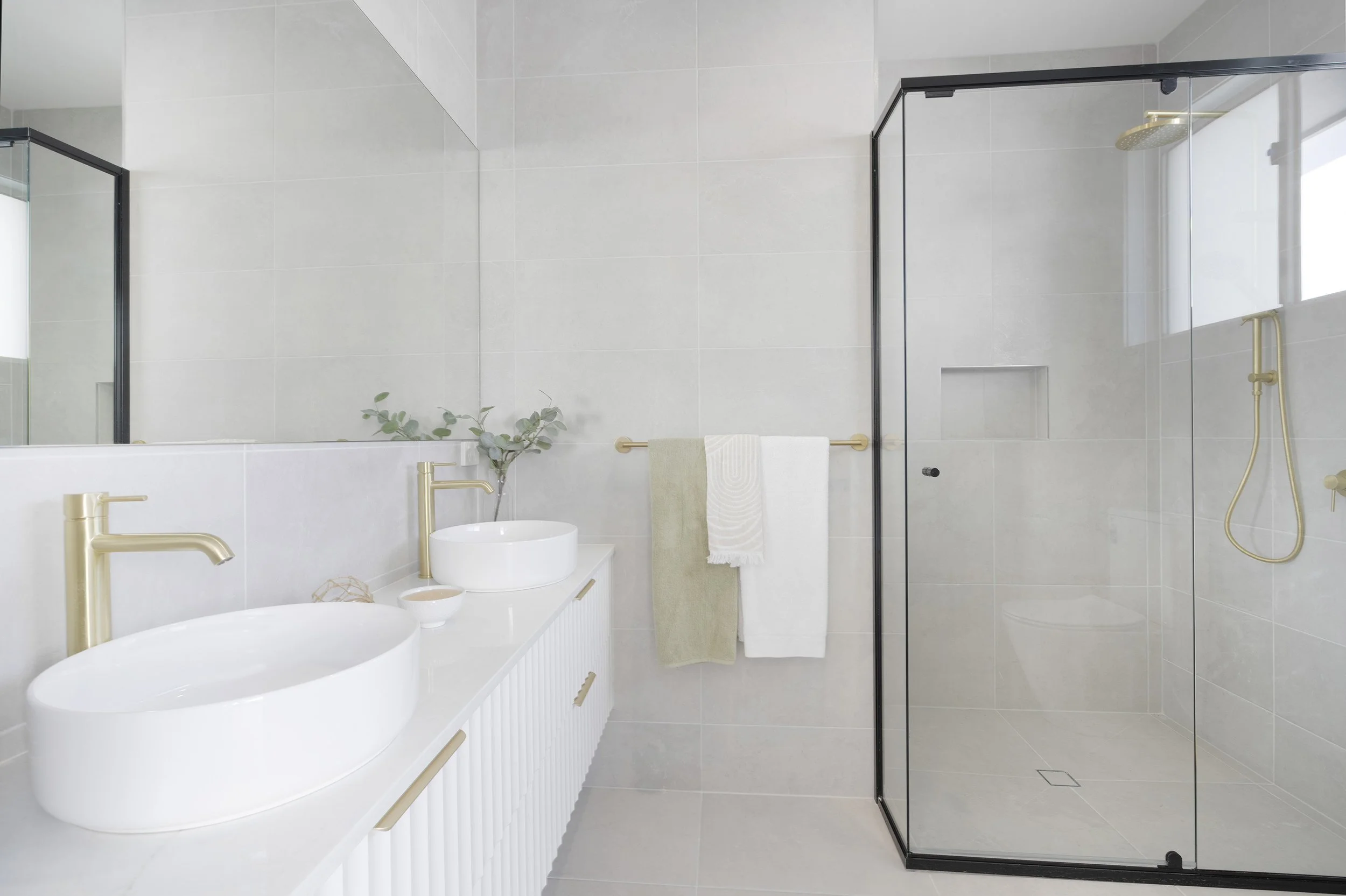Modern minimalist bathroom with white walls and tiles, gold fixtures, a glass shower enclosure, and a vanity with two vessel sinks.