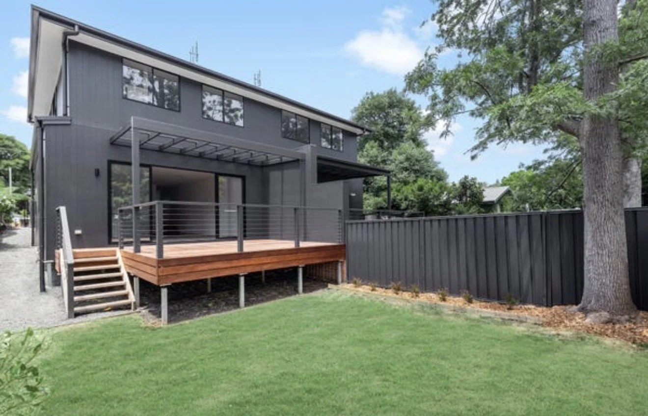 A modern two-story house with dark exterior walls, large windows, and a wooden deck in a yard