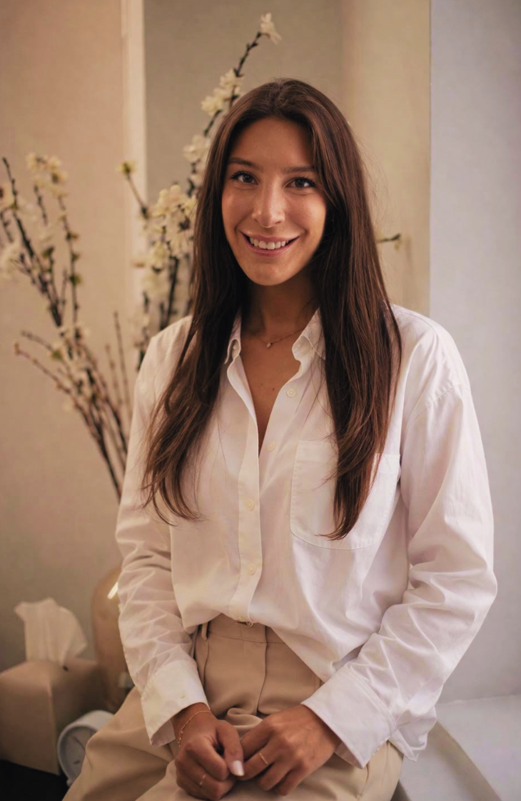 A woman with long brown hair and a white button-up shirt sitting in front of a beige wall and a vase with white flowers.