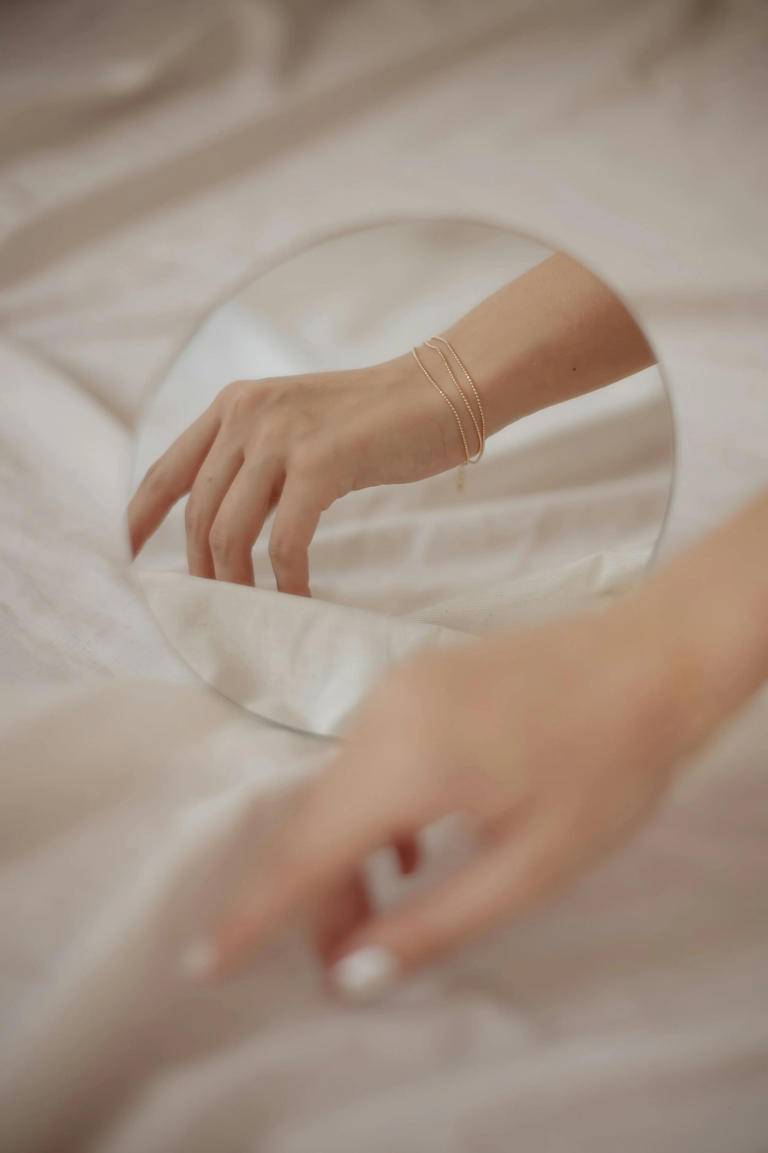 A hand with three thin gold bracelets held in front of a mirror with a silky white fabric background.
