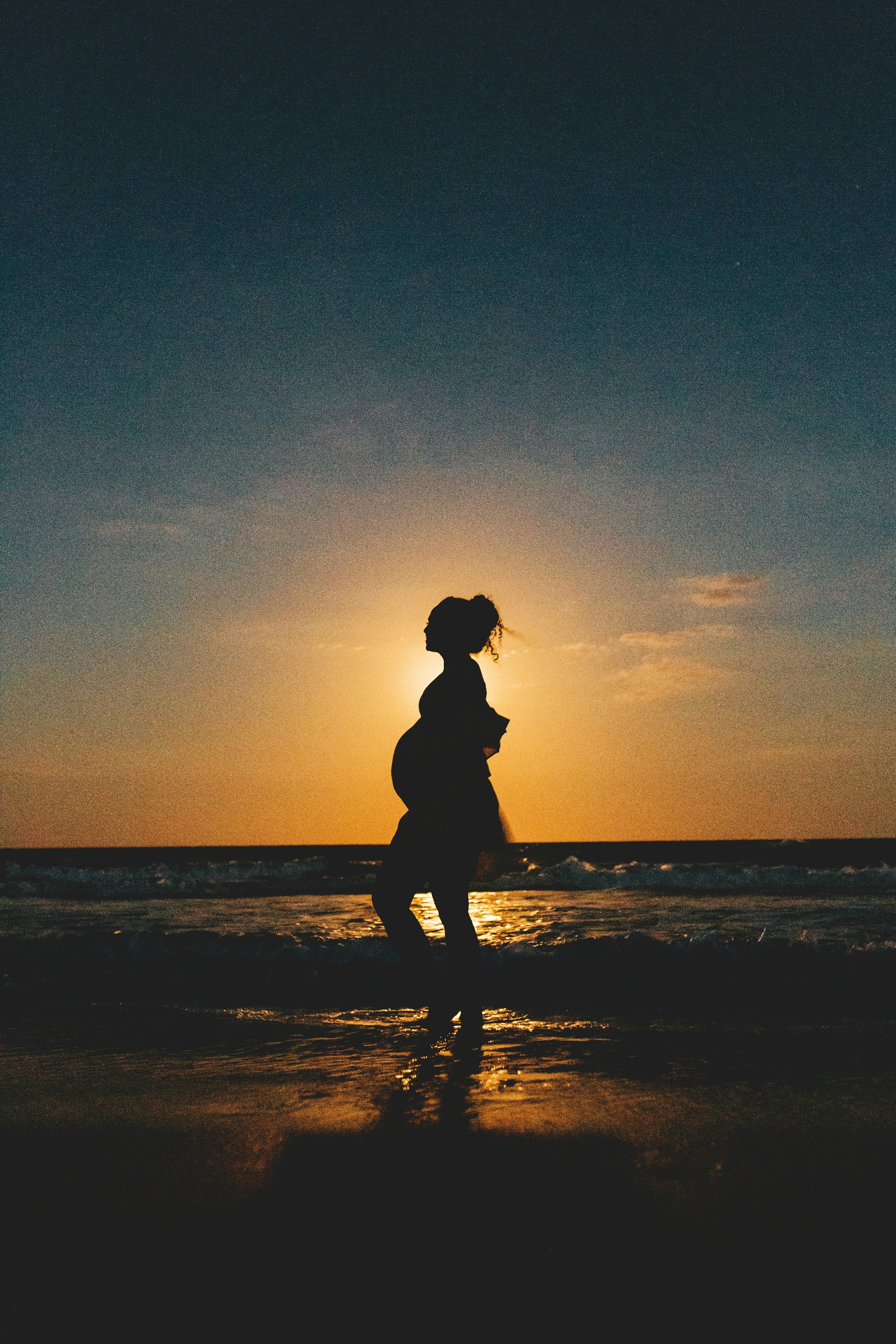 Silhouette of a woman with curly hair standing on the beach at sunset, with the sun setting behind her, creating a warm glow and reflecting on the water.