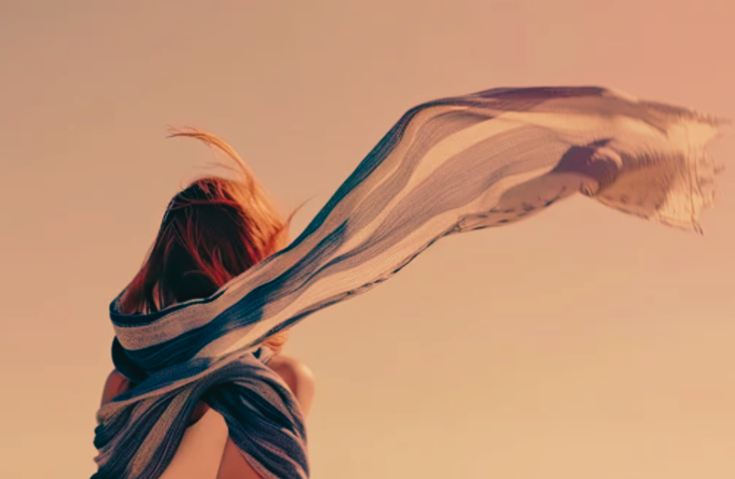 A person with hair blowing in the wind, wrapped in a large striped scarf, against a clear sky.
