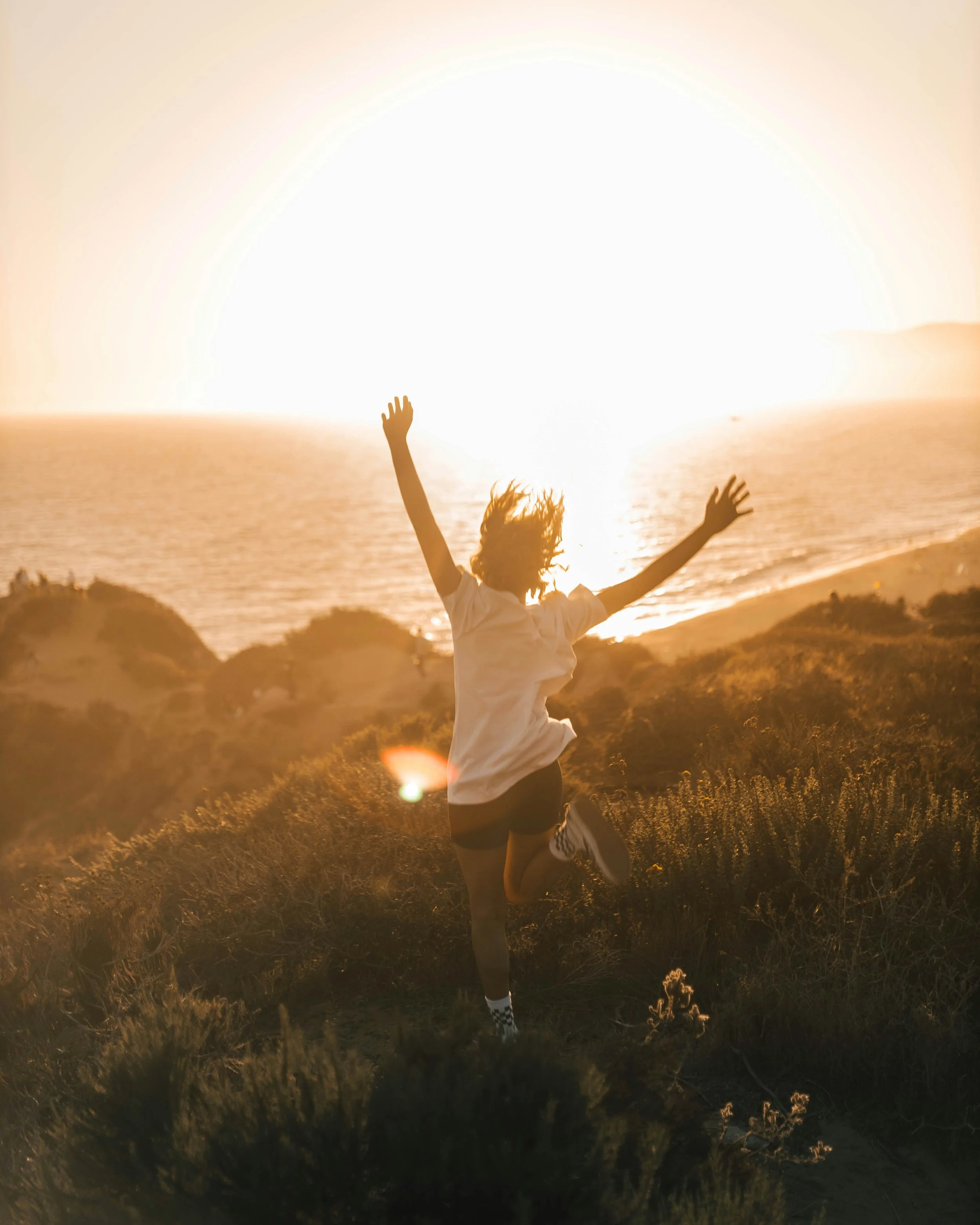 A person running on a grassy field at sunset with arms raised and hair flowing, with the sun setting over the ocean in the background.