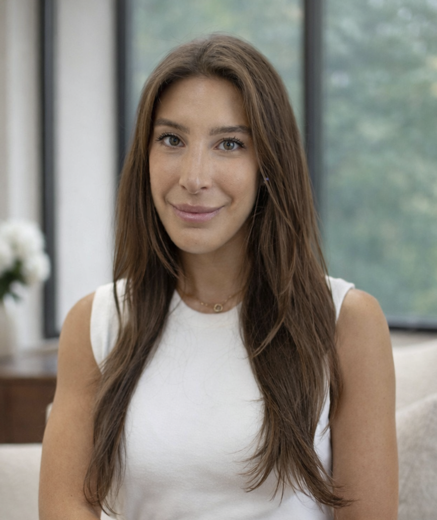 A young woman with long brown hair, wearing a white sleeveless top, sitting indoors with large windows and green trees in the background, smiling softly at the camera.