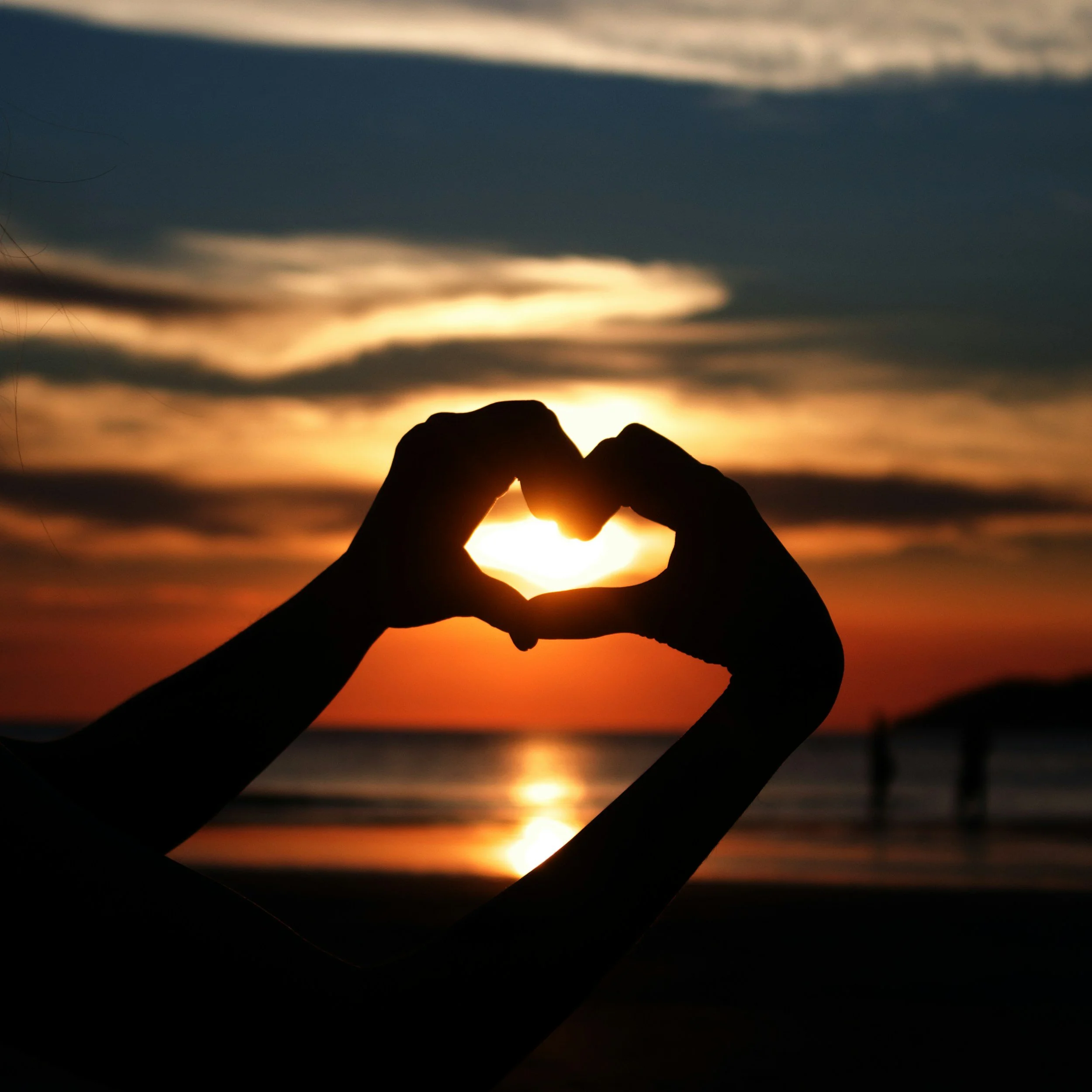 Silhouette of hands forming a heart shape against the sunset over the ocean with clouds in the sky.