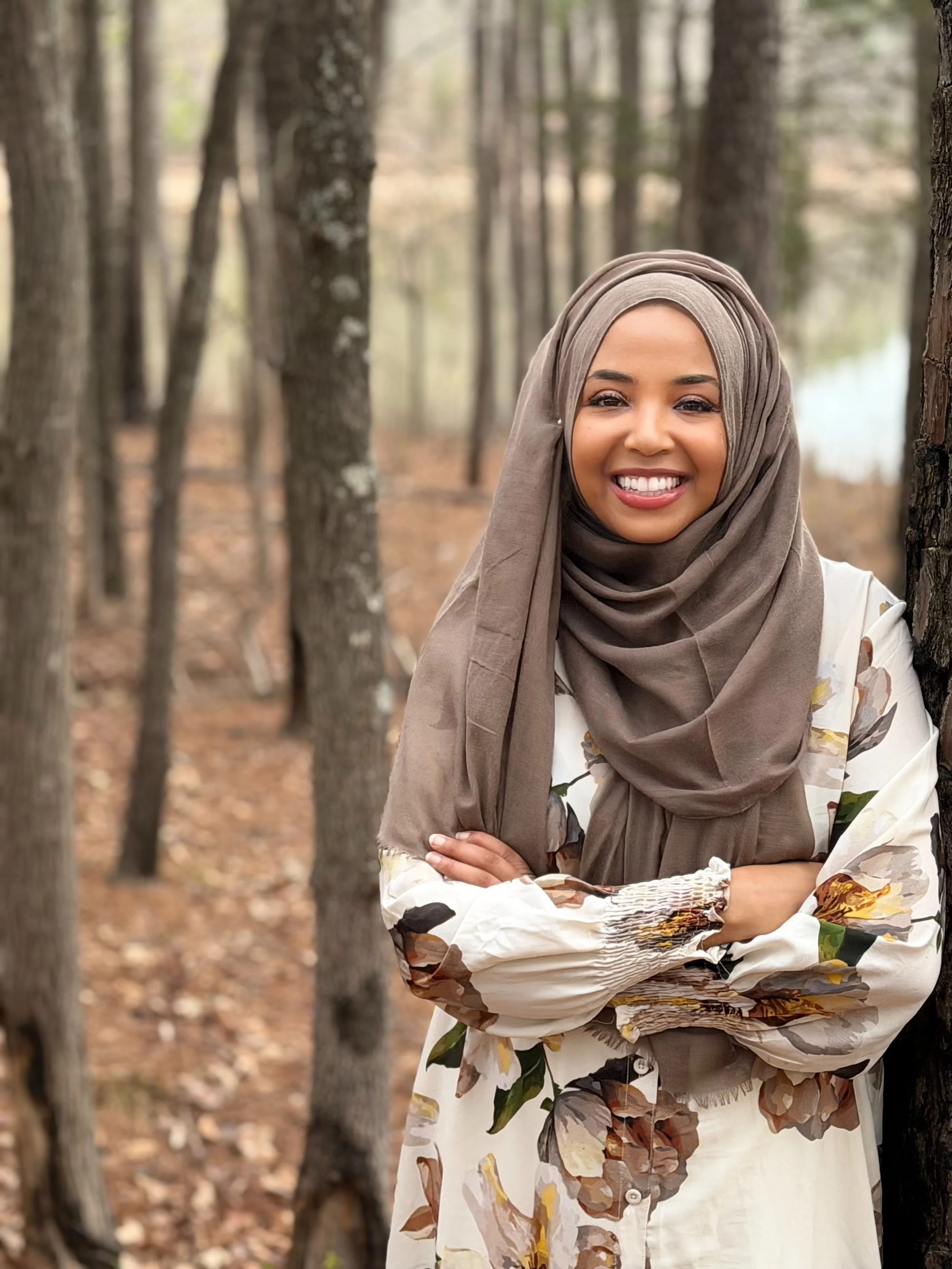 A woman wearing a beige hijab and a floral-patterned dress, standing in a forest with tall trees and fallen leaves on the ground, smiling and crossing her arms.