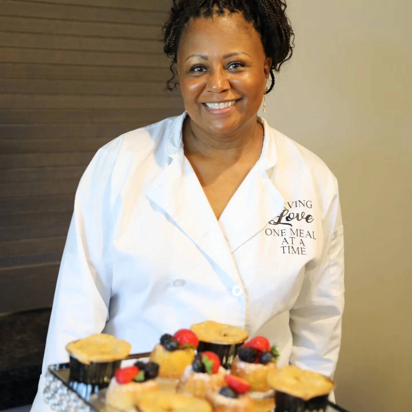 Woman smiling in a white chef's coat with embroidered words, standing behind a tray of assorted desserts topped with berries.