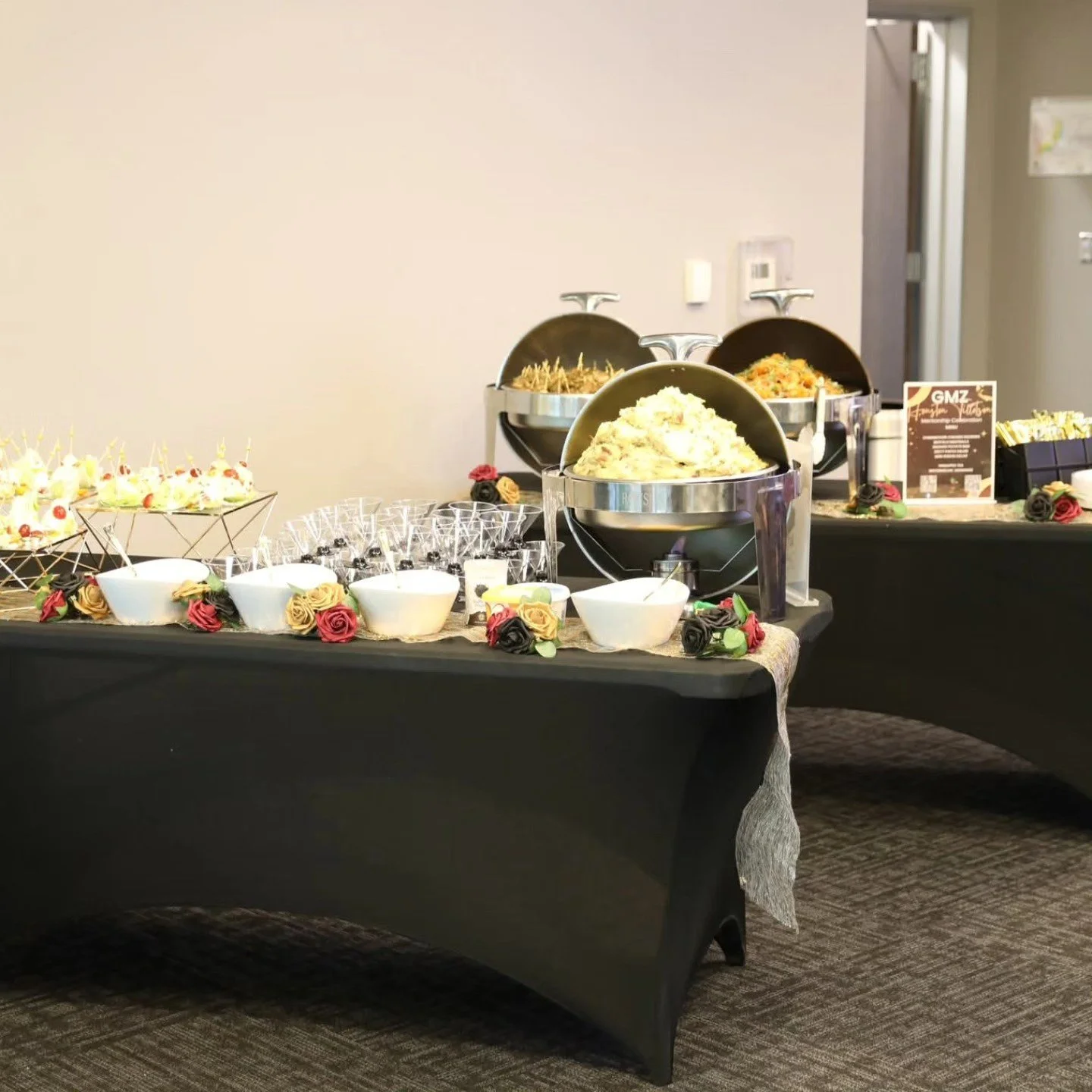 Buffet table with bowls of potato salad, fruit skewers, and various hot dishes, decorated with artificial roses, set up for an event.