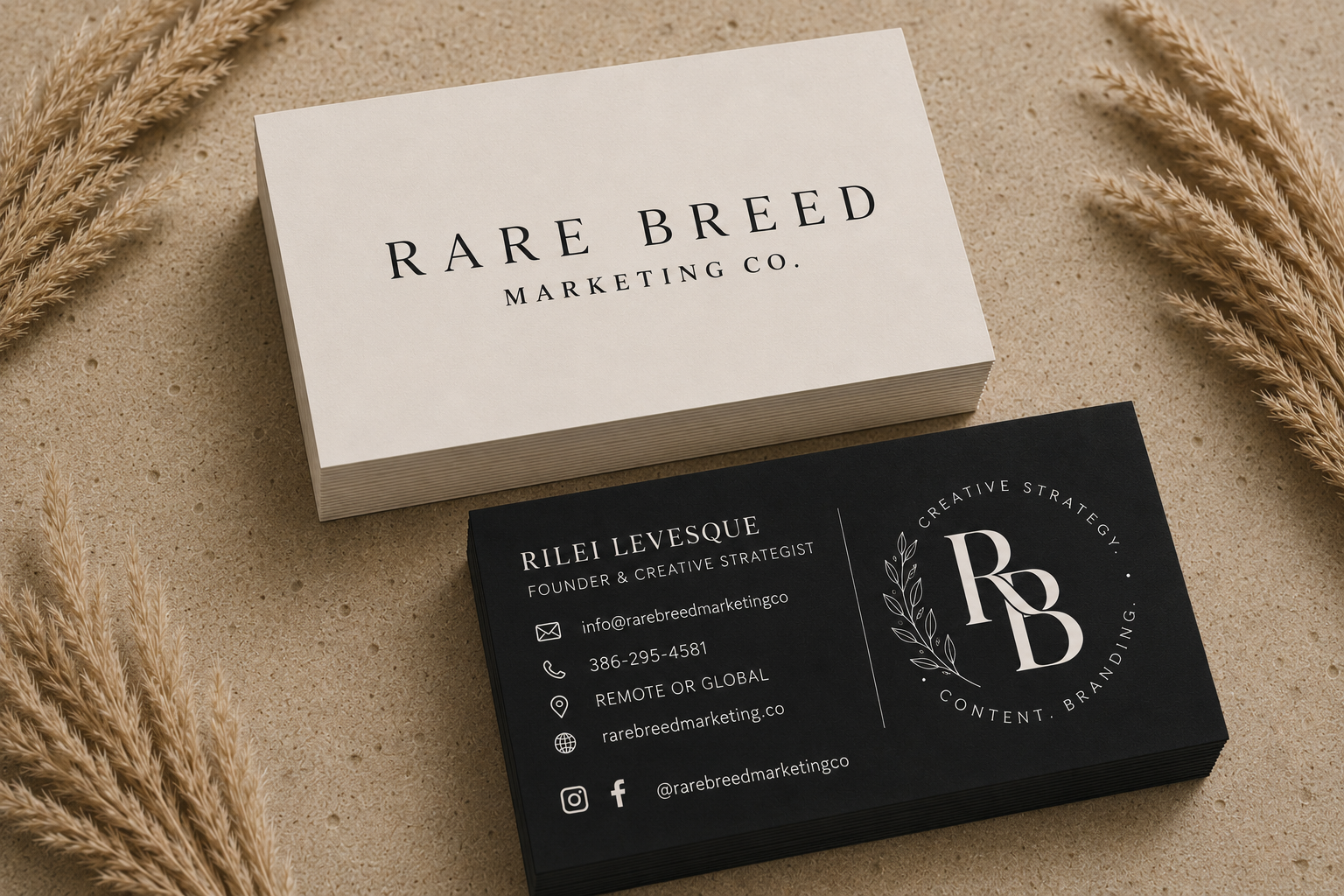 Business cards for Rare Breed Marketing Co., one black and one white, placed on a sandy surface with dried plants around.