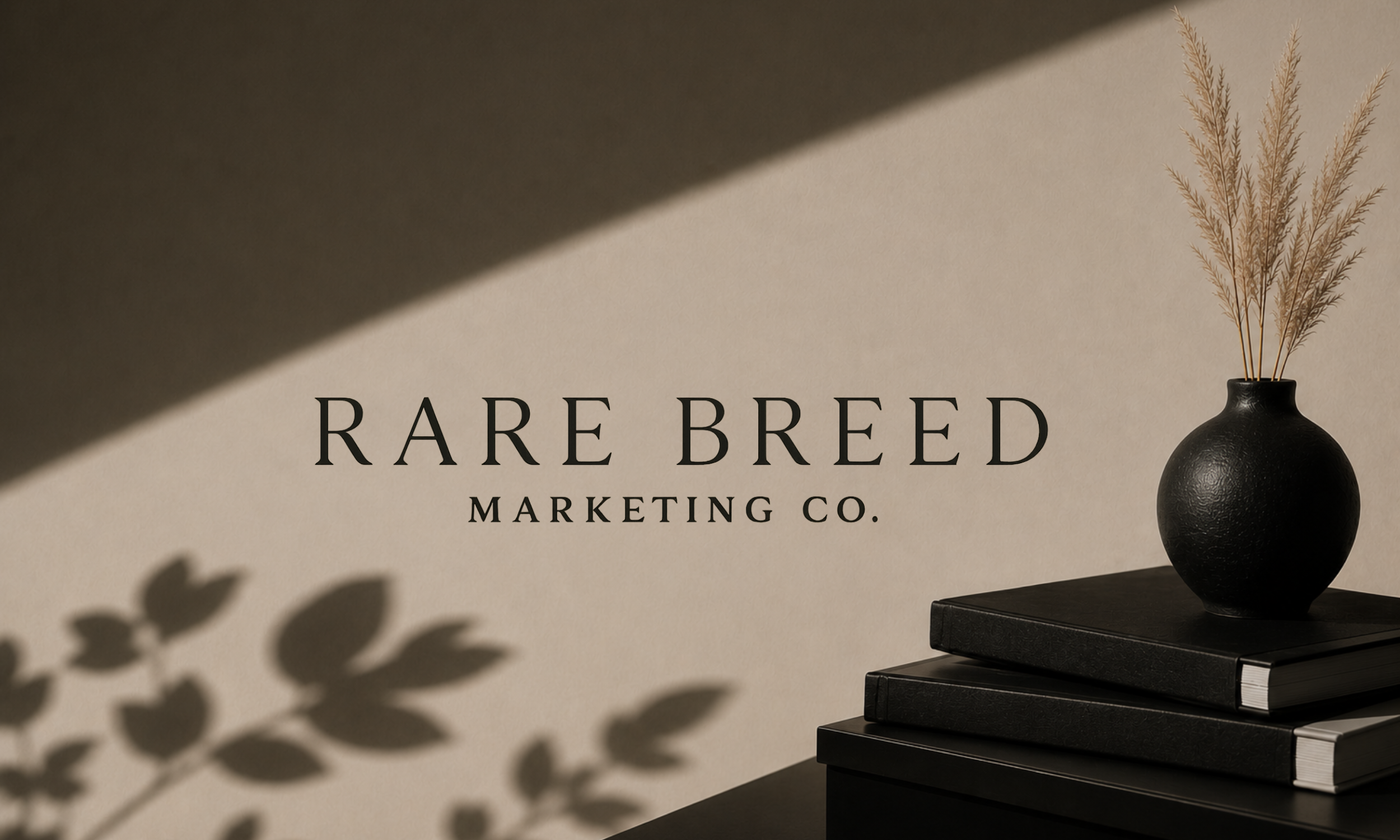 A black vase with dried beige pampas grass sits on a black table, with a shadow of leaves cast on a beige wall behind it, featuring the text 'RARE BREED MARKETING CO.' in black capital letters.