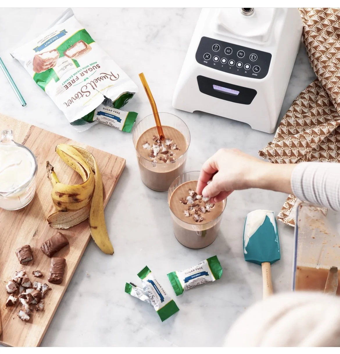 Two glasses of chocolate smoothies topped with shredded coconut, with a hand placing some coconut on top. On the table, there are bananas, a pack of sugar-free coconut, chocolate pieces, a cutting board, a mixer machine, a spatula, a small bowl, and a sugar-free coconut wrapper. The scene appears to be a kitchen counter preparing smoothies.