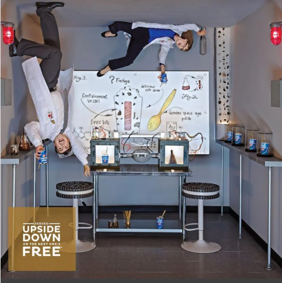 Two people in a science lab room, one upside down and one sideways. They are holding containers of yogurt, next to a whiteboard with drawings and notes about a science experiment involving Oreo cookies and fudge. There are science equipment, tables, and stools in the room.