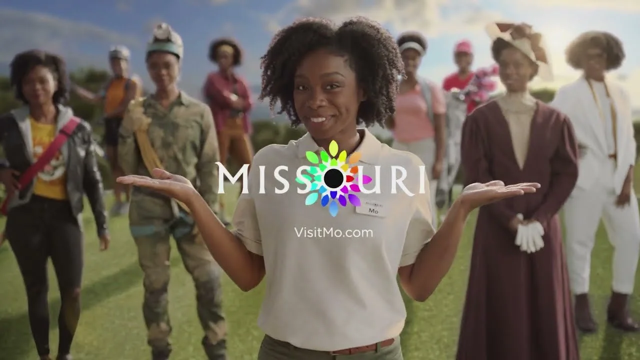 A group of diverse women standing outdoors, with a woman in the foreground smiling and holding her hands up, promoting Missouri with the VisitMo.com logo.