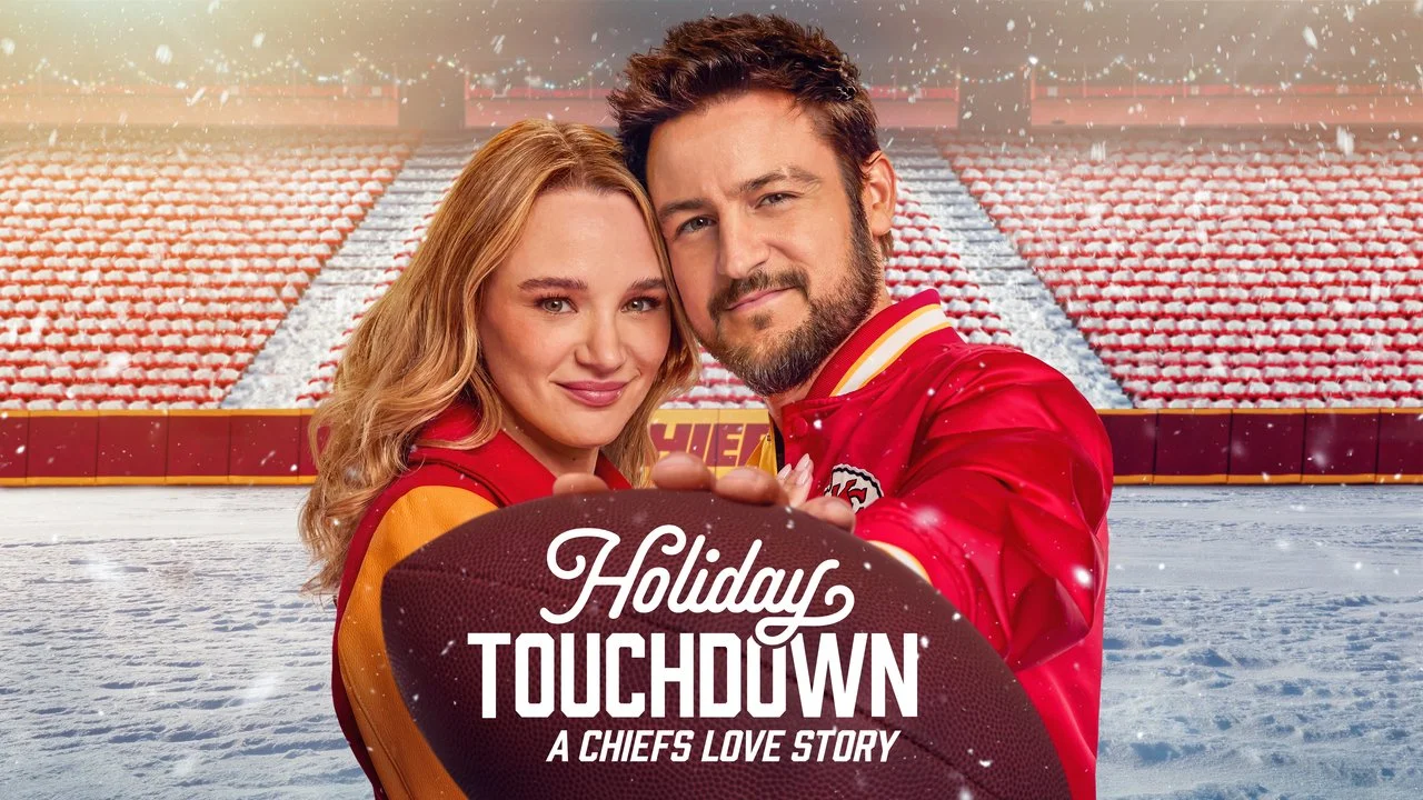 A man and woman in Chiefs jackets holding a football on a snowy football field with stadium seats in the background, promoting the movie 'Holiday Touchdown: A Chiefs Love Story.'