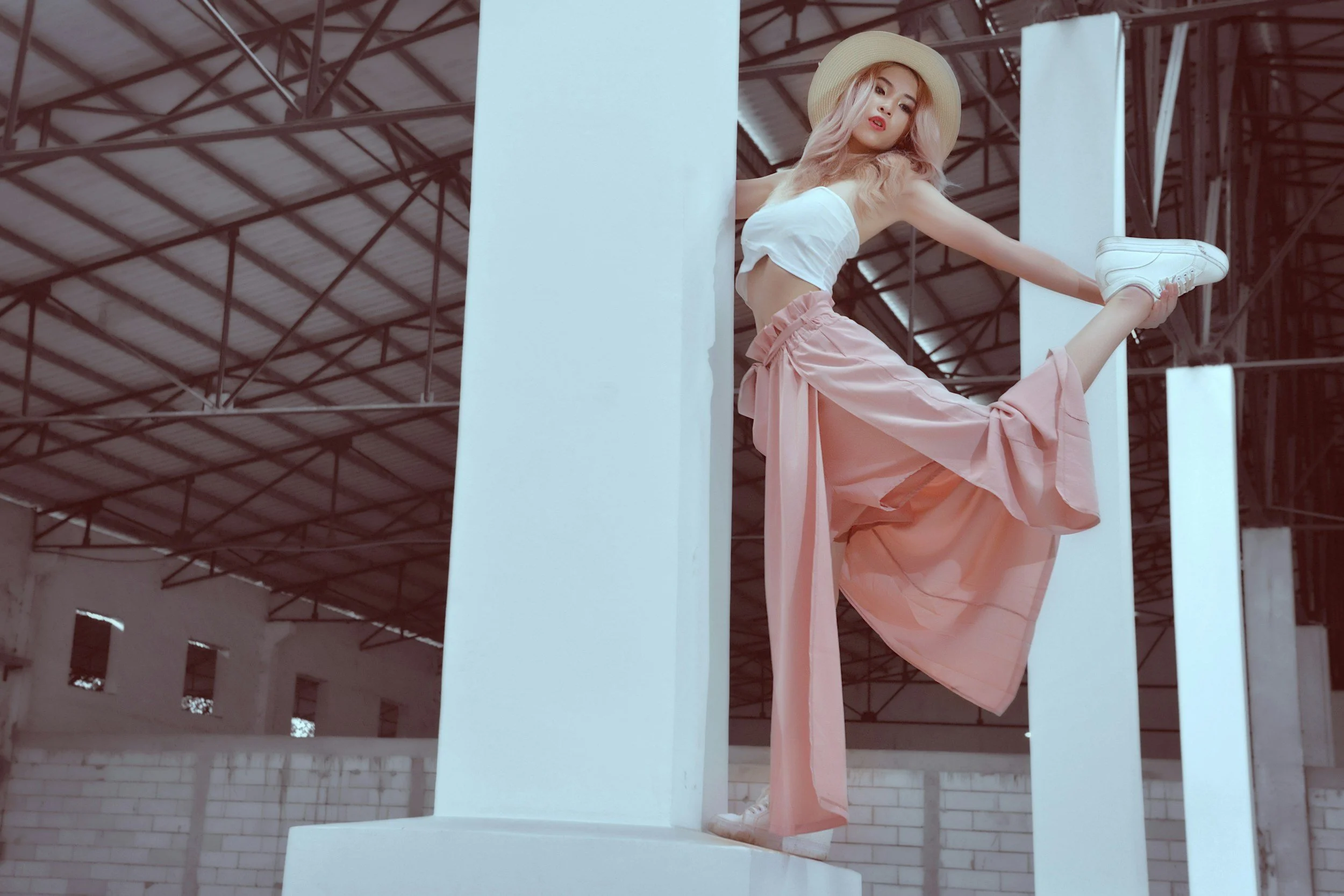 A woman with pink hair wearing a wide-brimmed hat, white crop top, and pink wide-leg pants with sneakers, poses on a white structure inside a large industrial warehouse.