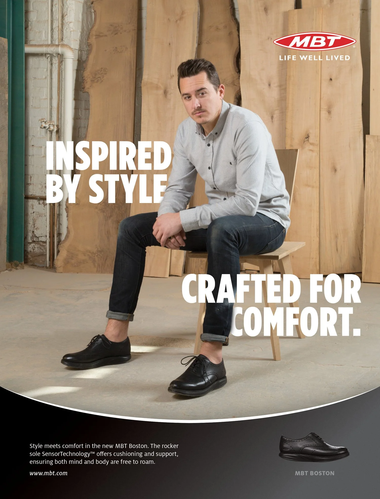A man sitting on a wooden chair in front of a wooden background, wearing a light gray button-up shirt and black shoes. Overlaid text reads 'Inspired by Style crafted for comfort' and a logo for MBT with the slogan 'Life Well Lived'.