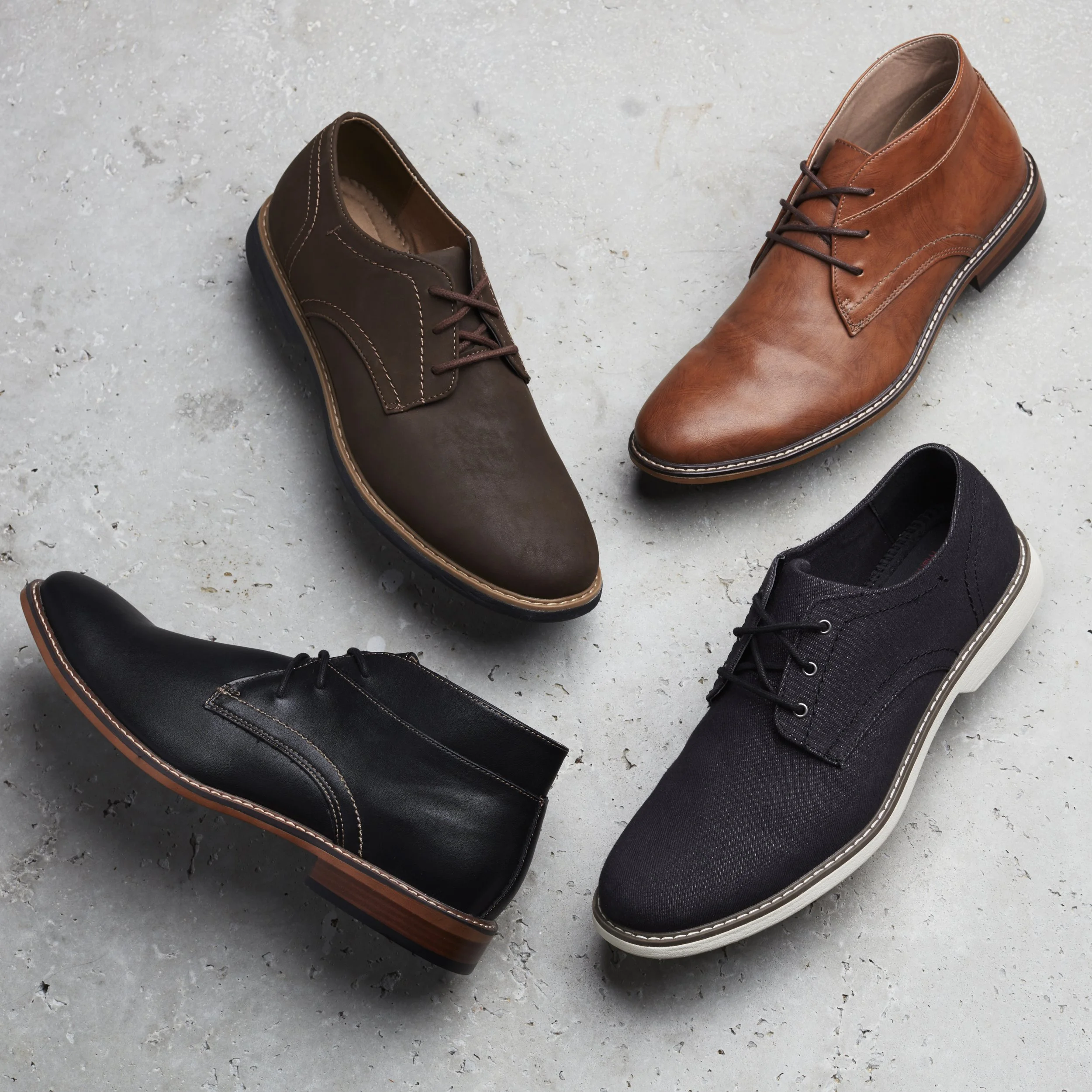 Three pairs of men's shoes arranged on a concrete floor, including a black pair with white soles, a brown pair with white soles, and a tan pair with darker soles.