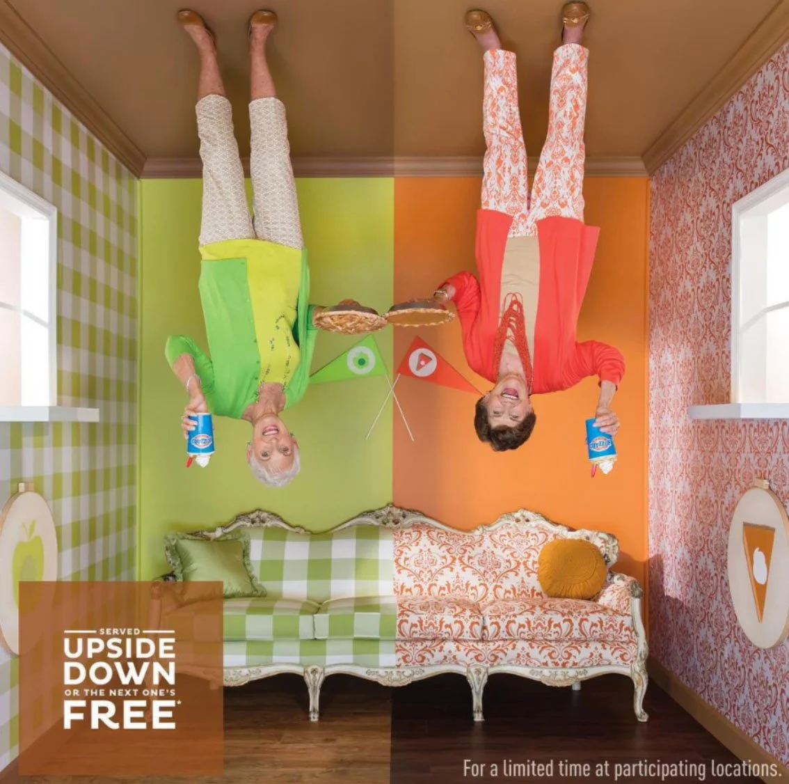 Upside-down room with two women holding pizza slices and soda bottles, smiling, with colorful plaid and patterned wallpaper, and a vintage-style sofa.