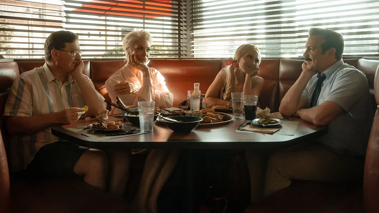 Four people sitting around a dining table with food and drinks in a restaurant with blinds and sunlight.