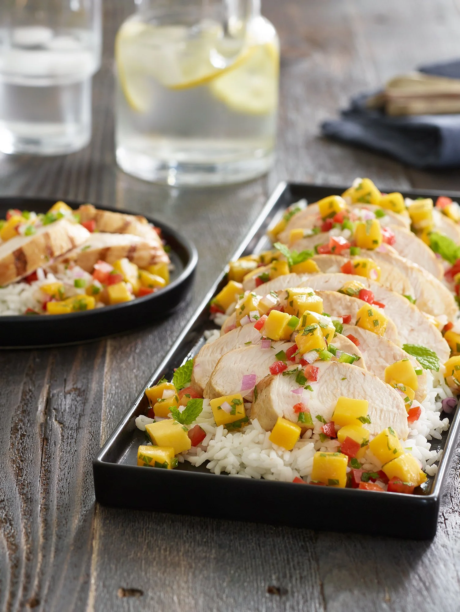 Sliced chicken breast topped with mango, red bell pepper, onion, and cilantro, served over rice with lime wedges and water on a rustic wooden table.