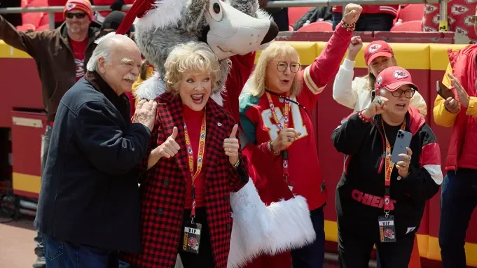 Fans at a sports game celebrating, with some wearing team merchandise, taking photos, and a mascot in the background.