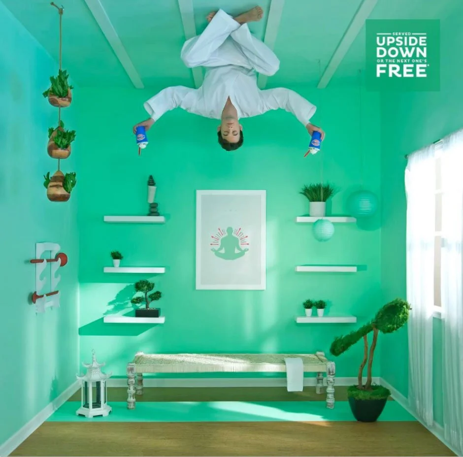 A person in white clothing depression upside down on the ceiling holding two cups of tea or coffee, with a green room decorated with potted plants, shelves, a bench, and decorative items, designed for a yoga or wellness space.