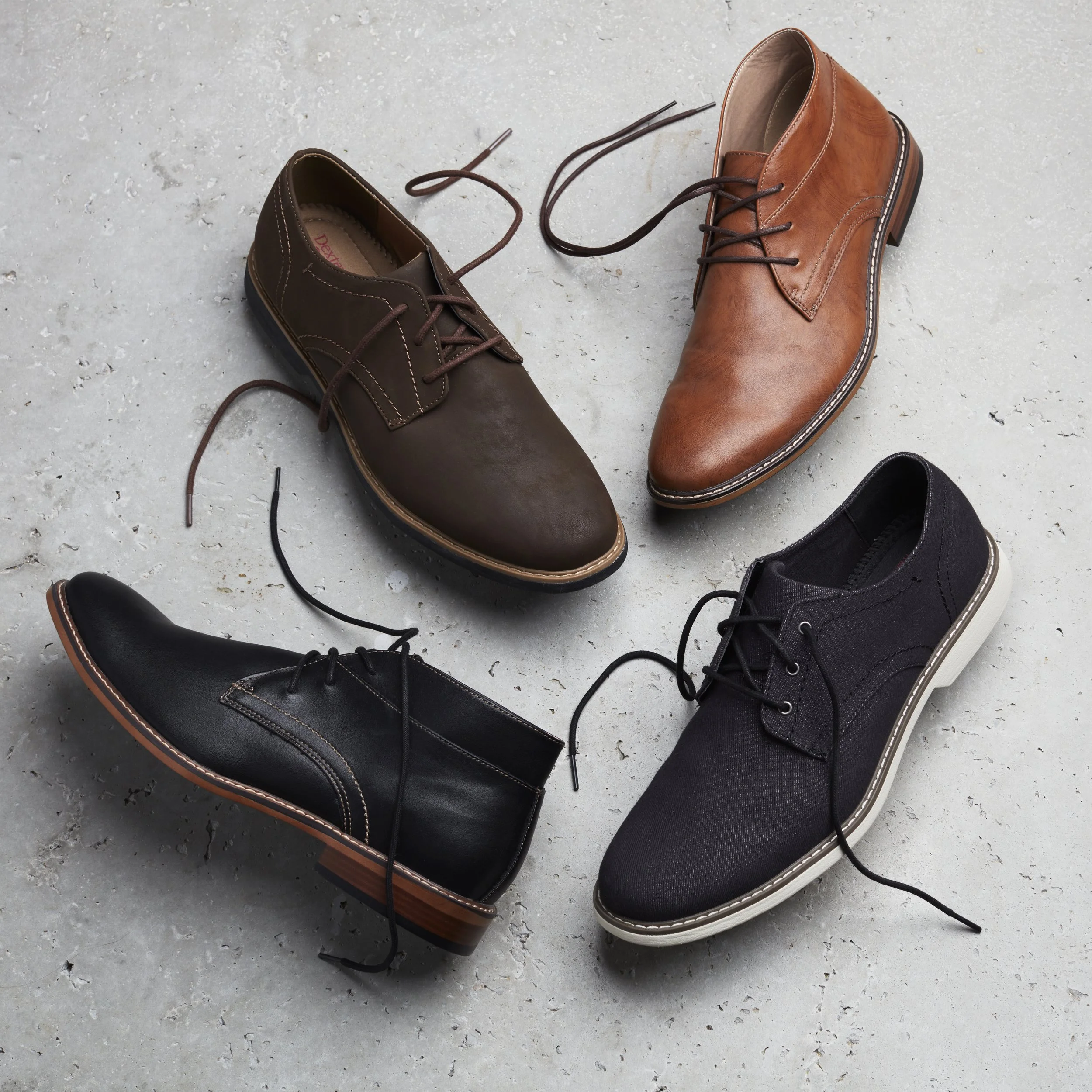 Four different pairs of shoes arranged in a circle on a concrete surface. The pairs include black, brown, light brown, and black shoes, with shoelaces tied.