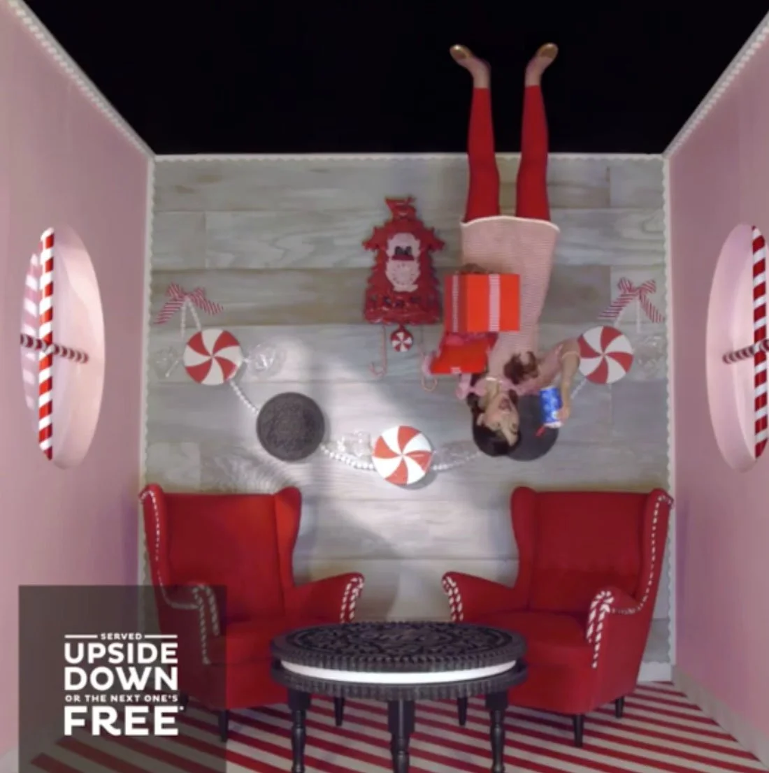 Upside-down Christmas-themed room with pink and red decor, including red chairs with white trim, striped red and white rug, candy cane decorations, peppermint swirls, a Christmas tree, a girl in red tights and a skirt, and a woman with a blue cup.